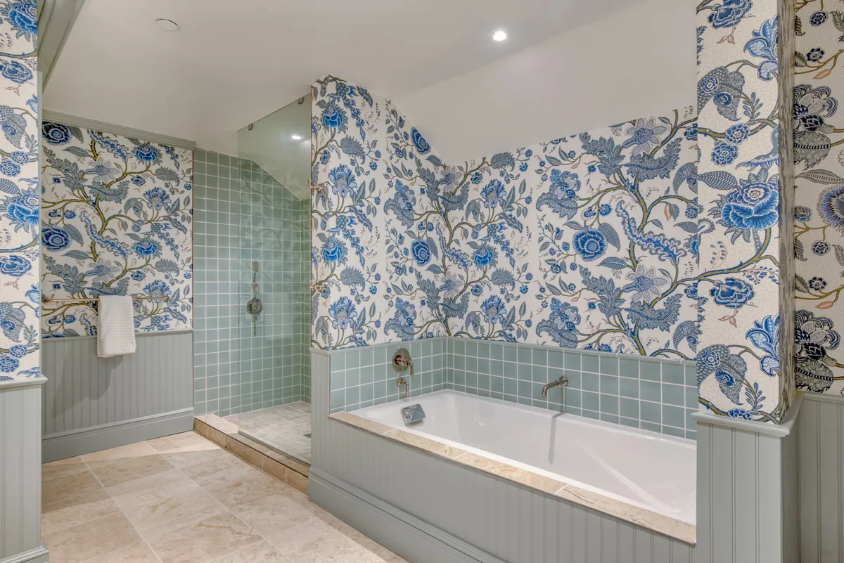 The image showcases a serene, spa-like bathroom featuring a luxurious freestanding bathtub, a glass-enclosed shower, and elegant floral wallpaper. The space is bathed in soft natural light, creating a tranquil atmosphere perfect for unwinding after a day at the beach.