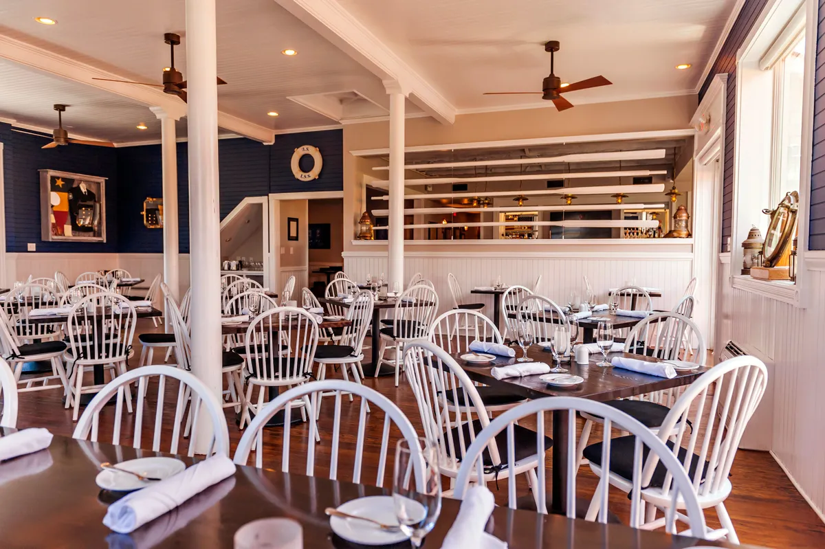 A bright, welcoming dining space featuring white wooden chairs, dark wooden tables, decorative wall art, and an open kitchen. The ambiance is casual yet stylish, making it ideal for healthy, communal meals and social gatherings.