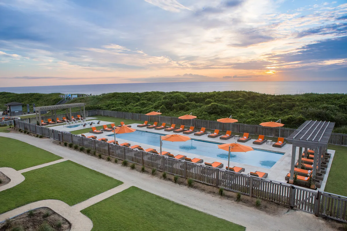 A serene poolside scene with lounge chairs and orange umbrellas facing the ocean. The tranquil water and scenic backdrop create an inviting space for sunset gatherings or quiet moments by the pool.