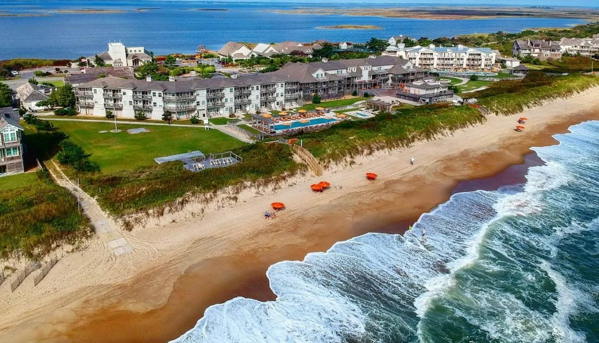 A sweeping exterior view of the resort's beachfront, combining the inviting hotel architecture, sparkling swimming pool, and umbrellas dotting the sandy beach. Families can be imagined sunbathing, swimming, or playing along the water’s edge, all set against the picturesque backdrop of the ocean.