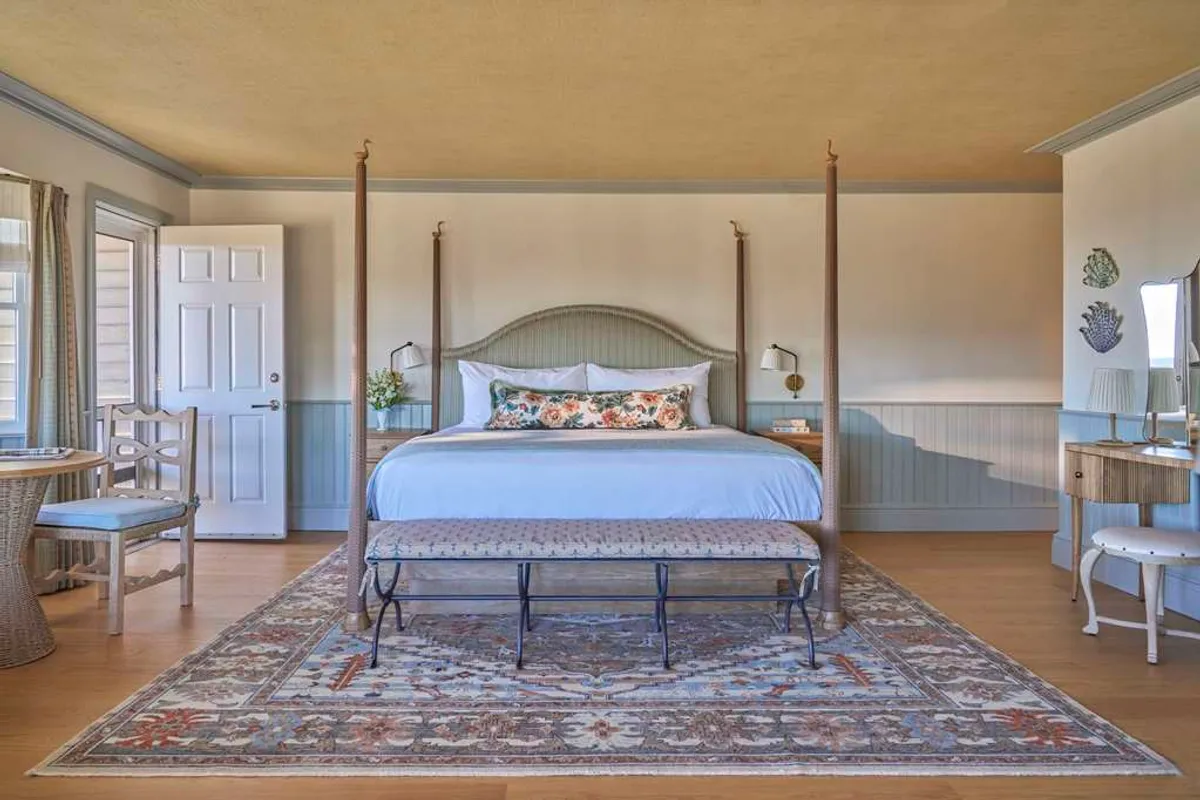 A tranquil bedroom featuring a four-poster king bed with crisp linens, a decorative rug, a vanity desk, and a cozy seating area by the window. Morning light gently illuminates the room, enhancing its serene atmosphere and highlighting thoughtful details that encourage relaxation.