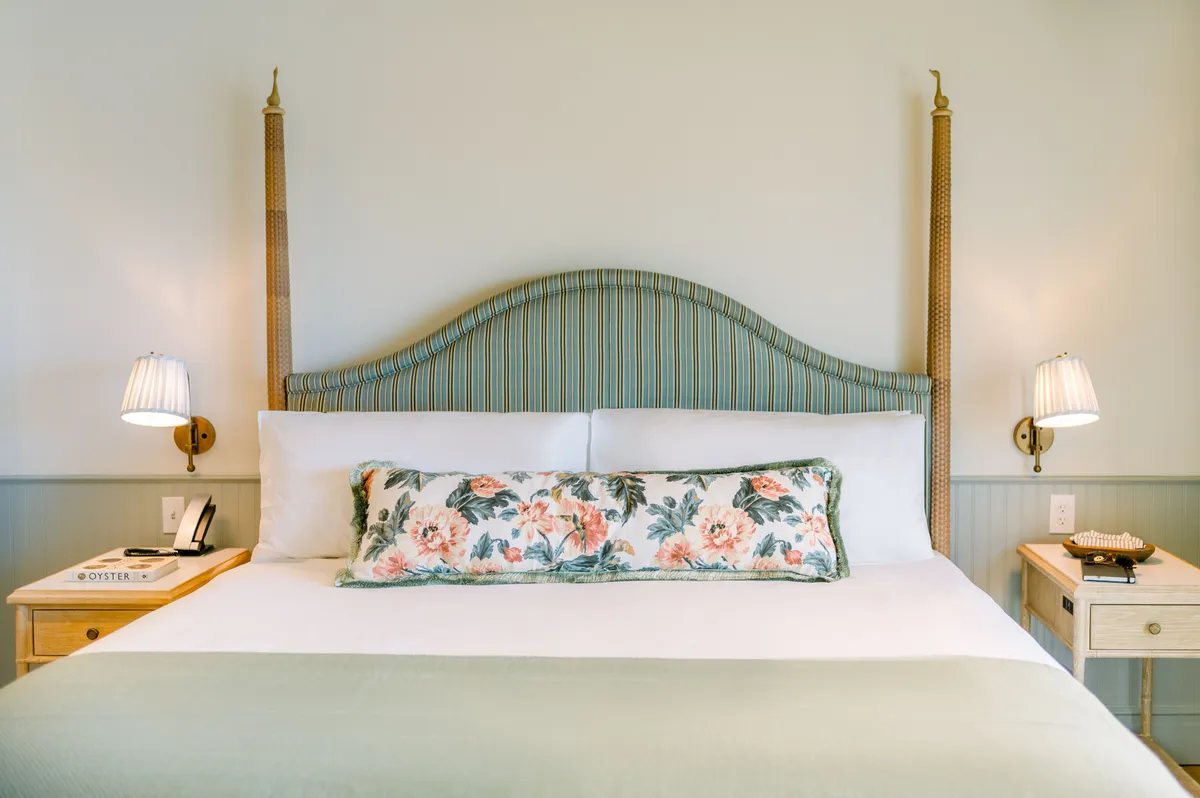 A cozy guest room filled with natural light, featuring a plush bed with a high headboard, colorful floral pillows, and a softly glowing lamp on a nightstand. Decorative items add a touch of personality and comfort, evoking the inviting ambiance of a relaxing retreat.