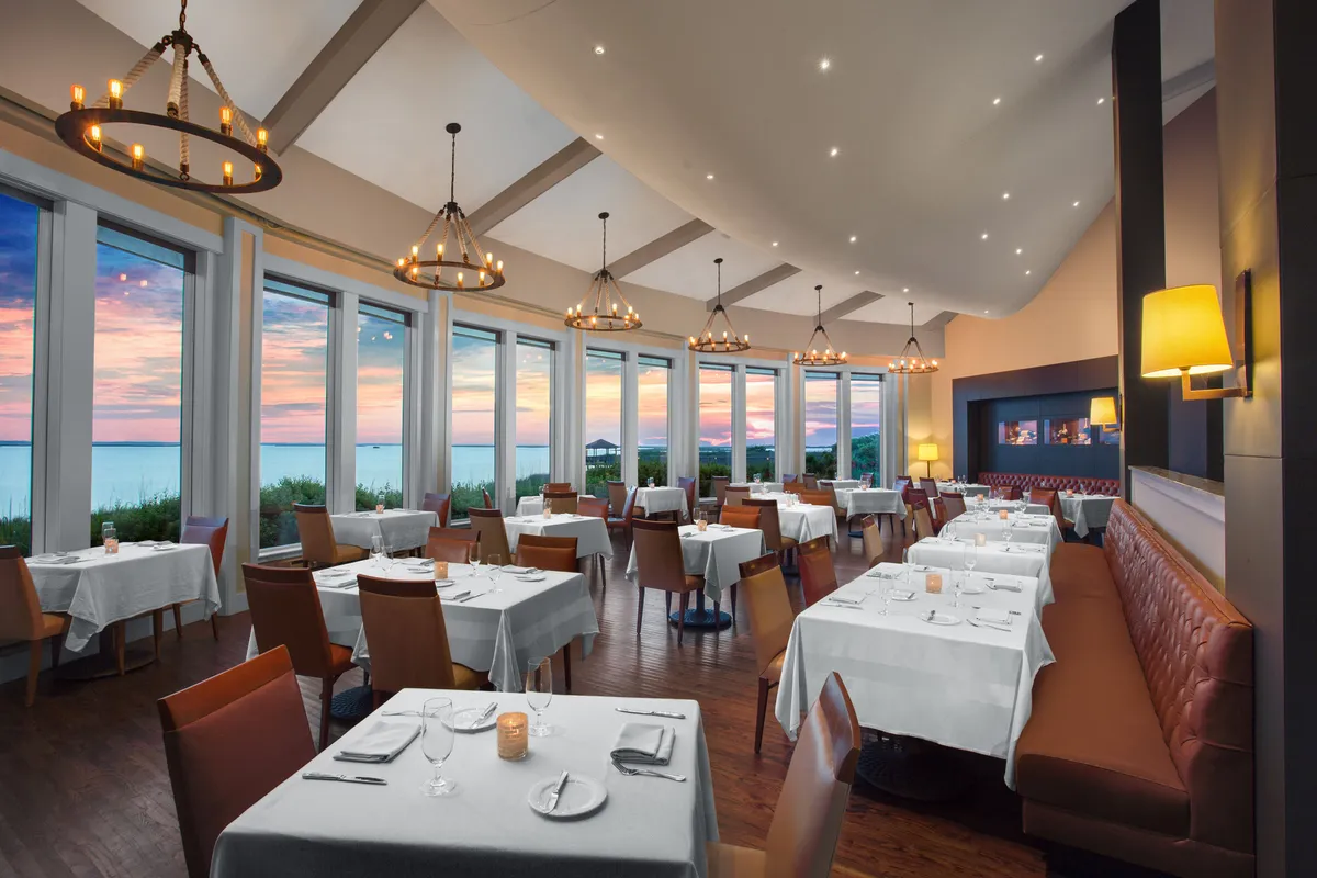 An elegant dining room with large windows opening to a sunset view, ambient lighting from chandeliers, and beautifully set tables ready for a memorable meal.