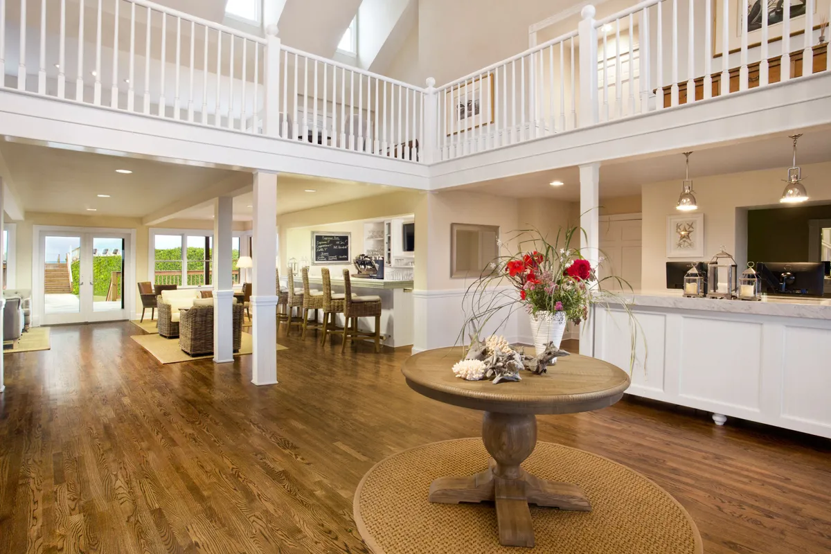 This image features a round dining table with a floral arrangement, an open kitchen area, and large windows that let in ample natural light. The high ceilings with exposed beams create a spacious, welcoming atmosphere, and the open layout seamlessly connects the dining and living spaces.