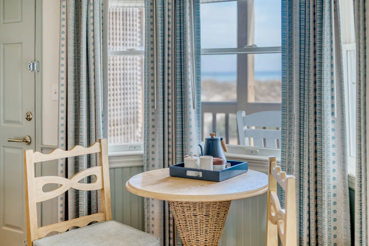 A cozy interior scene featuring a wooden table and woven chair positioned near airy curtains, with a tea set ready for use. The scene suggests a welcoming space to enjoy a morning coffee, read a book, or share an intimate conversation. Light streaming through the window hints at a view just beyond, enhancing the sense of a relaxing retreat.