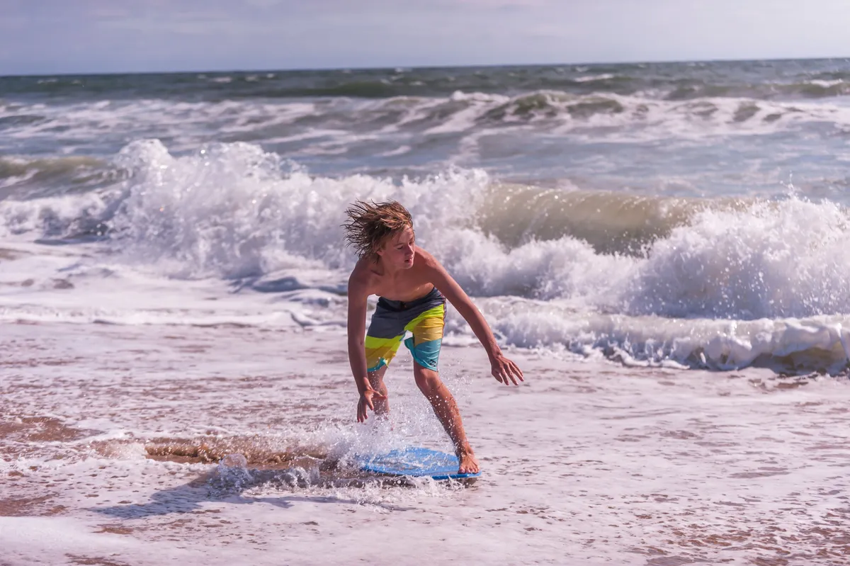 A joyful scene of a young boy with a boogie board running along the sandy shoreline, with waves splashing in the background and the vast ocean stretching beyond. The activity and the vibrant beach environment evoke a classic family beach day filled with play and adventure.