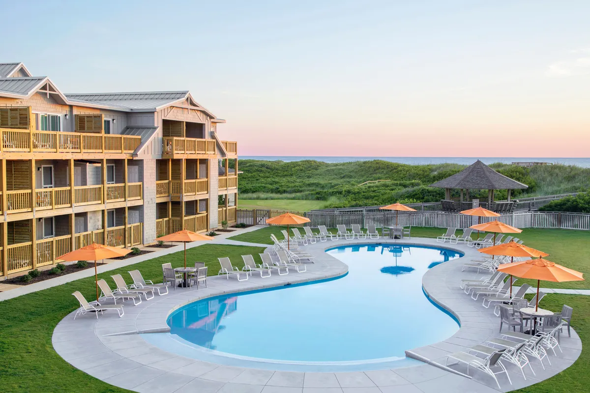 A sparkling outdoor swimming pool surrounded by elegant lounge chairs and large umbrellas, with the resort building's facade and lush landscaping in the background. The scene is sunlit, inviting guests to swim, sunbathe, or relax poolside.