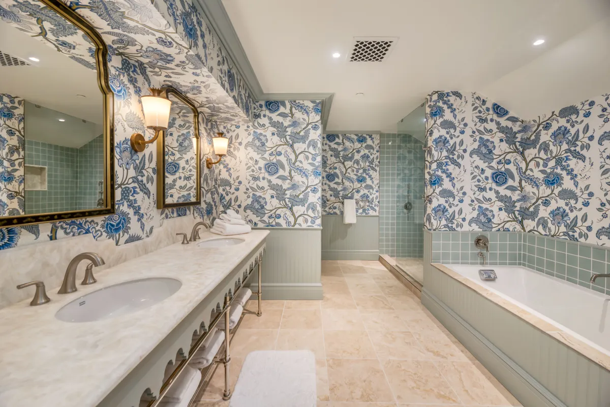 A sophisticated bathroom scene featuring ornate mirrors, floral wallpaper, a marble countertop, and a bathtub set against a window with a calming view. The setting is serene and elegant, ideal for unwinding after a long day.