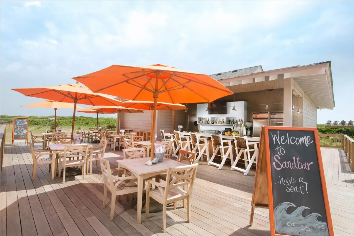 An open-air dining area is depicted, with bright orange umbrellas shading wooden tables and chairs. Guests can enjoy drinks or dinner in a cheery, laid-back environment while taking in the fresh air and coastal ambiance.