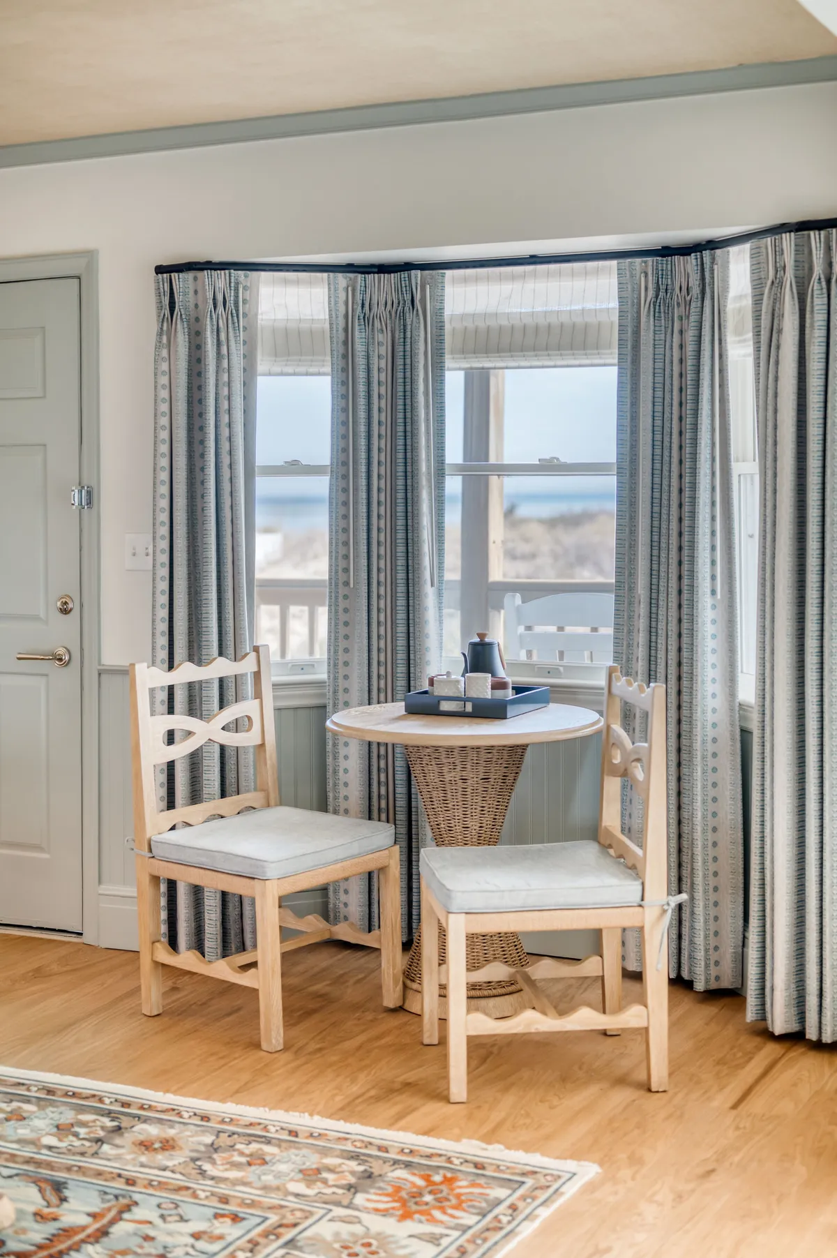 A cozy round table with two wooden chairs sits beside curtained windows, offering a view of the outdoors. The soft, natural light and inviting setup suggest a perfect spot for coffee, reading, or intimate conversation.