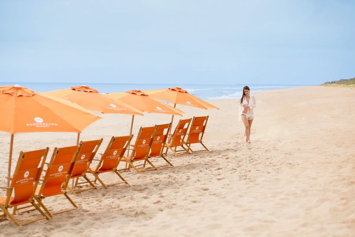 An inviting exterior scene shows beach chairs and umbrellas facing the ocean, with a person strolling along the sandy shore. The calm, open setting underscores panoramic views and leisure, suggesting the type of vista available from a private balcony.