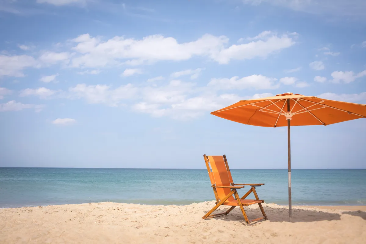 A bright orange beach chair and umbrella set on soft sand, overlooking clear blue water. The beach is calm and inviting, perfect for sunbathing, reading, or simply taking in the ocean views.