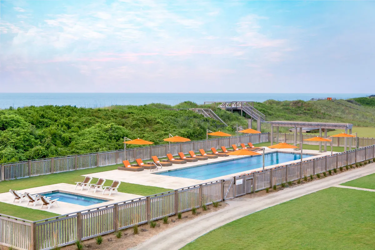 A serene outdoor swimming pool area, surrounded by lush green landscaping, with inviting lounge chairs and orange umbrellas. The scene conveys an atmosphere ideal for relaxation, socializing, and enjoying wellness-focused activities like swimming or yoga by the poolside.