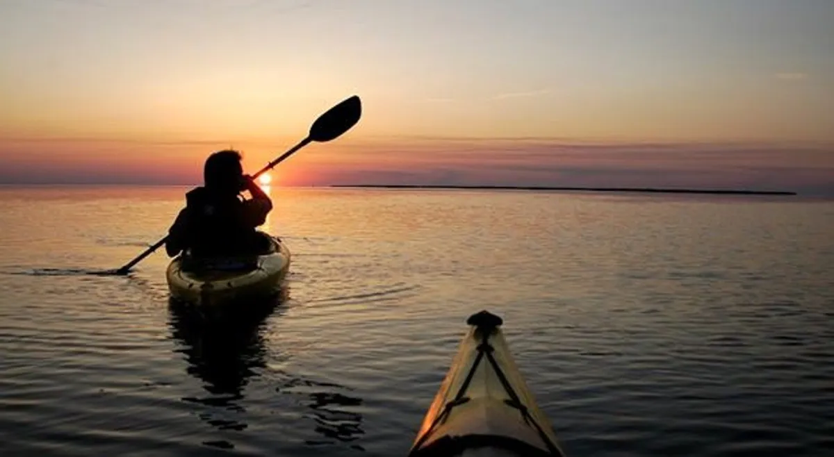 A kayaker paddles peacefully on still water at sunset, with the sky aglow in warm hues and the tranquil scene reflecting on the water’s surface. The image evokes the spirit of outdoor adventure and serene exploration unique to the area.